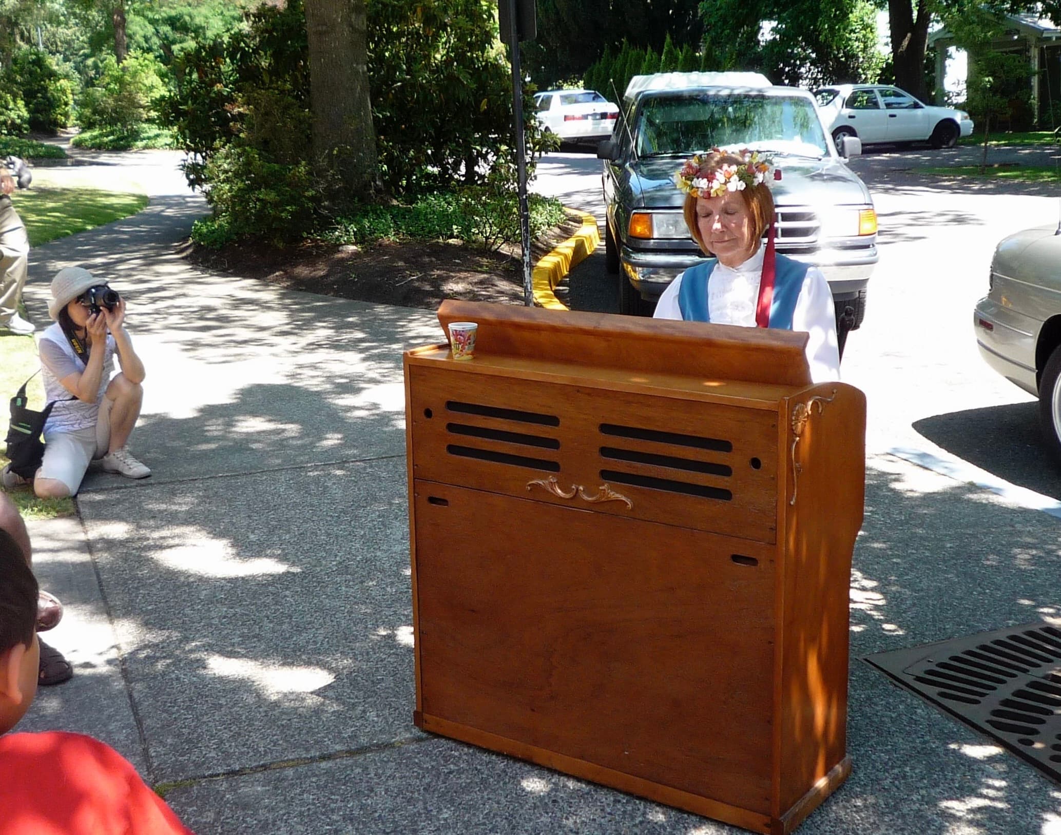 Wanda in a flower crown playing a wooden pump organ outdoors while someone photographs her